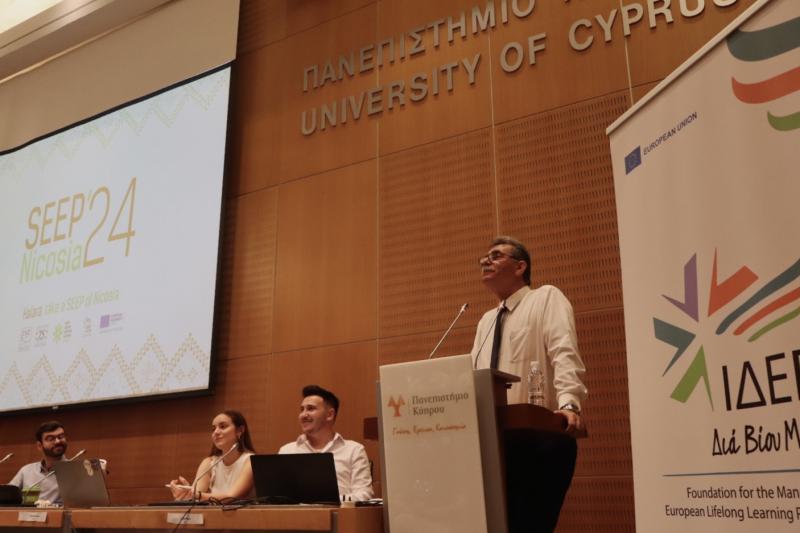 A panel discussion, in which there is a speaker delivering a speech, a long desk of other speakers in the back, and a big projection of the event's name