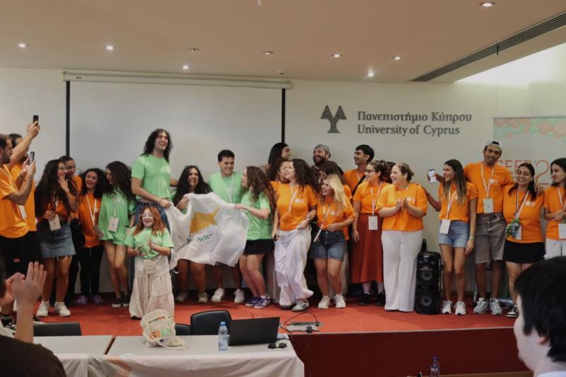 A group of volunteers dressed in orange t-shirts and holding a Cyprus flag, are cheering and jumping.