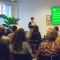 A photo of a person speaking in front of an audience.